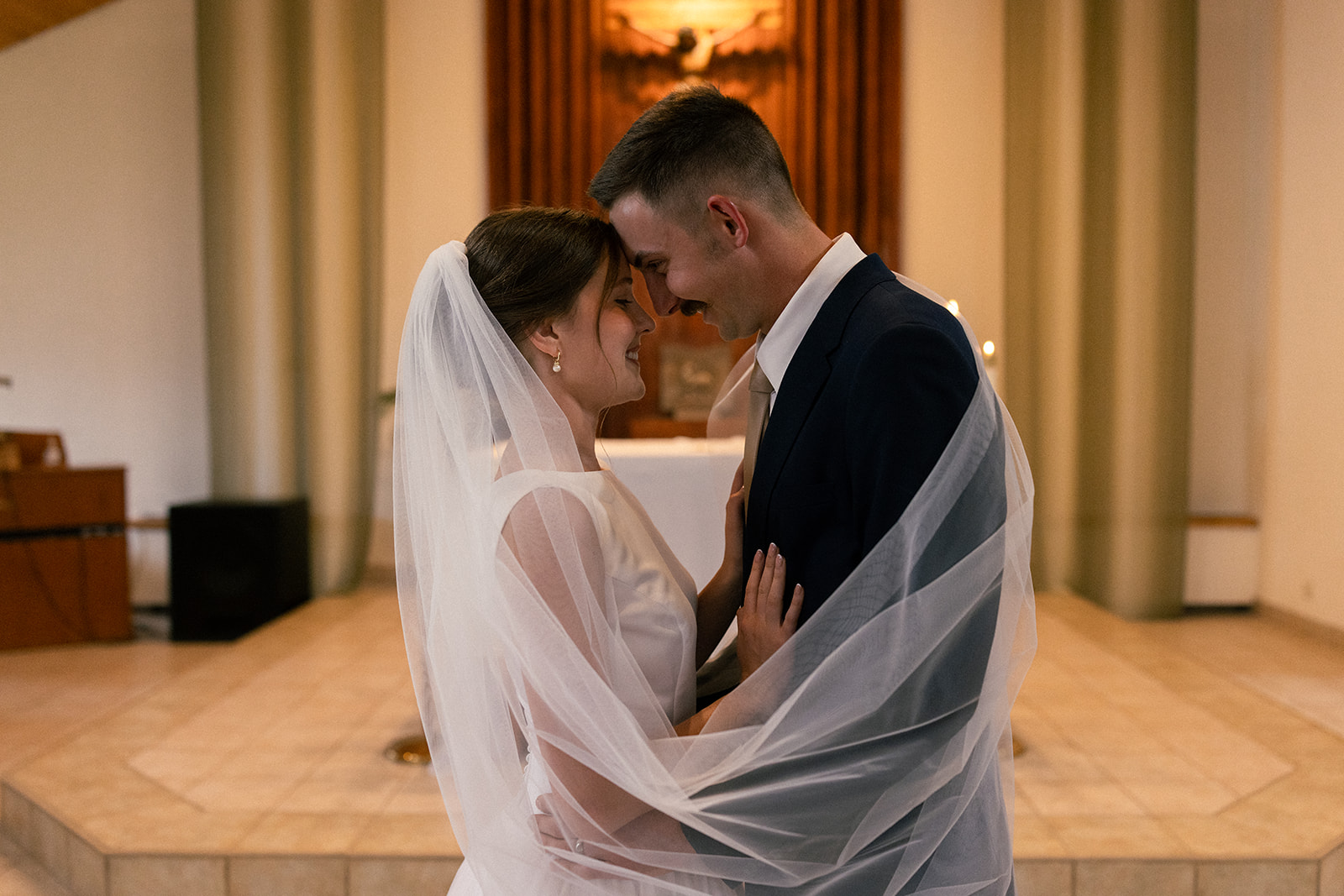 Bride and groom wrapped in a veil in front of the crucifix at St. John the Baptist Catholic church in Custer, SD taken by Catholic wedding photographer Alexis Dean